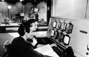 zwei Menschen arbeiten an einem Schaltpult in einem Fernsehstudio, im Hintergrund ein Kamerateam.