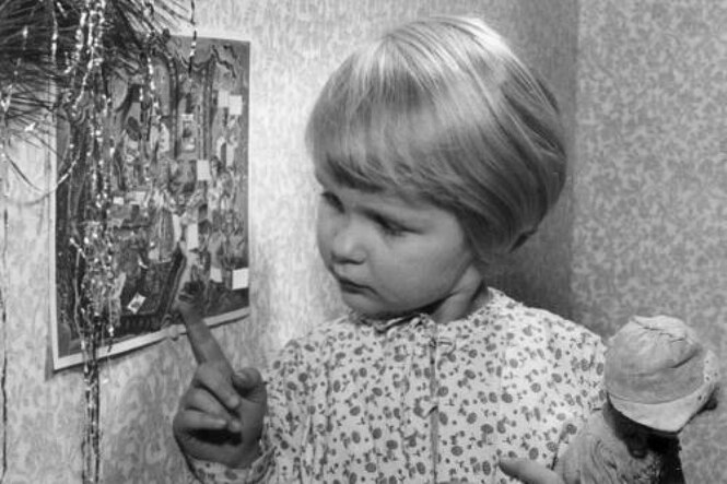 Ein kleines Mädchen im Nachthemd und Puppe im Arm schaut auf den Adventskalender, an dem schon mehrere Türchen geöffnet sind