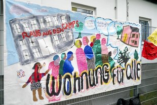 Das Bild zeigt ein selbst bemaltes Plakat. Darauf zu sehen sind mehrere Menschen, die in einer Schlange vor einem Haus anstehen. Darunter steht "Wohnung für alle" geschrieben.