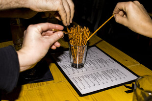 Drei Hände greifen nach Salzstangen, die in einem Glas auf einem gelben Tisch stehen