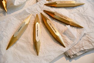 Goldene Trophäen des Reporter:innen-Preises in Bleistiftform mit Spitze je an beiden Enden. Sie liegen auf einem zerknitterten, weißen und edleren Papier.