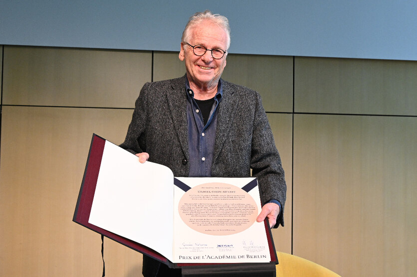 Daniel Cohn-Bendit, dunkles Jacket, schwarzes T-Shirt unter dem blauen Hemd, keine Krawatte hält lächlend die Urkunde des Preises in die Kamara