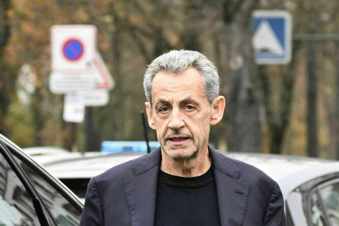 Nicolas Sarkozy in Shirt und Jacket steht zwischen Autos und schaut angestrengt