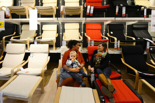 Zwei Frauen mittleren Alters sitzen entspannt in einem IKEA-Showroom auf roten und beigen Liegestühlen. Die eine hält ein Kleinkind auf dem Schoß, das seinen Schnuller inspiziert. Sie unterhalten sich, während im Hintergrund eine Wand voller weiterer IKEA-Stühle (helle Holzgestelle mit schwarzen, roten oder beigen Bezügen) aufgereiht ist, teilweise mit Preisschildern.
