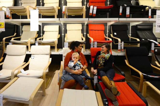 Zwei Frauen mittleren Alters sitzen entspannt in einem IKEA-Showroom auf roten und beigen Liegestühlen. Die eine hält ein Kleinkind auf dem Schoß, das seinen Schnuller inspiziert. Sie unterhalten sich, während im Hintergrund eine Wand voller weiterer IKEA-Stühle (helle Holzgestelle mit schwarzen, roten oder beigen Bezügen) aufgereiht ist, teilweise mit Preisschildern.