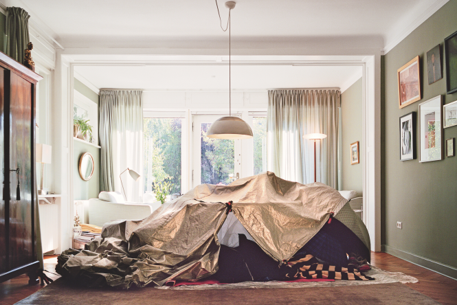 Ein Wohnzimmer mit stilvoll eingerichteter Altbau-Atmosphäre, helle Vorhänge und große Fenster, in der Mitte ein improvisiertes Zeltdach aus Decken und Folien aufgebaut wie ein provisorischer Unterschlupf; der Kontrast zwischen behaglicher Einrichtung und notdürftiger Lagerstruktur verweist auf das Thema Wohnungslosigkeit.