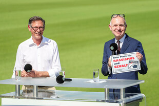 DFB-Präsident steht auf einem Rasenplatz vor einem Tisch mit Mikrophon und hält ein Schild hoch