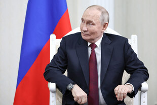 Wladimir Putin sitzt auf einem weissen Sessel, hinter ihm die russische Fahne, schaut nach links und lächelt versonnen