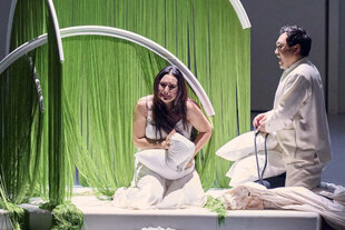 Zwei Schauspieler*innen in weiß knien während einer Darbietung vor aufgehängten grünen Vorhängen auf der Bühne und halten Kissen in den Armen.