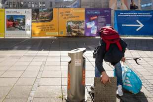 Ein Flaschensammler auf der Straße untersucht eine Bodenklappe neben einem Mülleimer, in der Hand eine halbvolle Tasche.