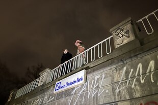 Zwei Personen hinter einer Brüstung. Unter ihnen ist an der Fassade ein Schild mit der Aufschrift 