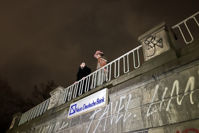 Zwei Personen hinter einer Brüstung. Unter ihnen ist an der Fassade ein Schild mit der Aufschrift "Neue Deutsche Bank" angebracht