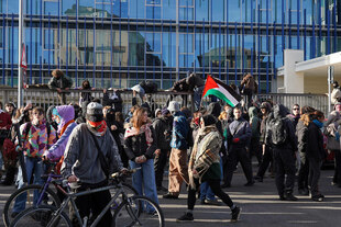 Eine Menschenmenge vor einem gläsernen Bürogebäude, einige tragen palestinensische Fahnen oder klettern über einen Zaun, der das Gebäude von der Straße trennt