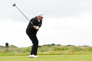 Donald Trump auf einem Golfplatz schwingt einen Golfschläger zum Abschlag. Er trägt dunkle Sportkleidung, eine weiße Baseballkappe und weiße Golfschuhe. Vor ihm liegt der Golfball auf einem Tee, im Hintergrund sind Grasflächen und hügelige Landschaft zu sehen, unter einem bewölkten Himmel.