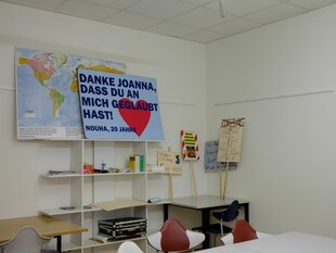 Ein Regal im Büroraum auf dem ein Schild steht mit der Aufschrift: "Danke Joanna, dass du an mich geglaubt hast. Nouha, 20 Jahre"