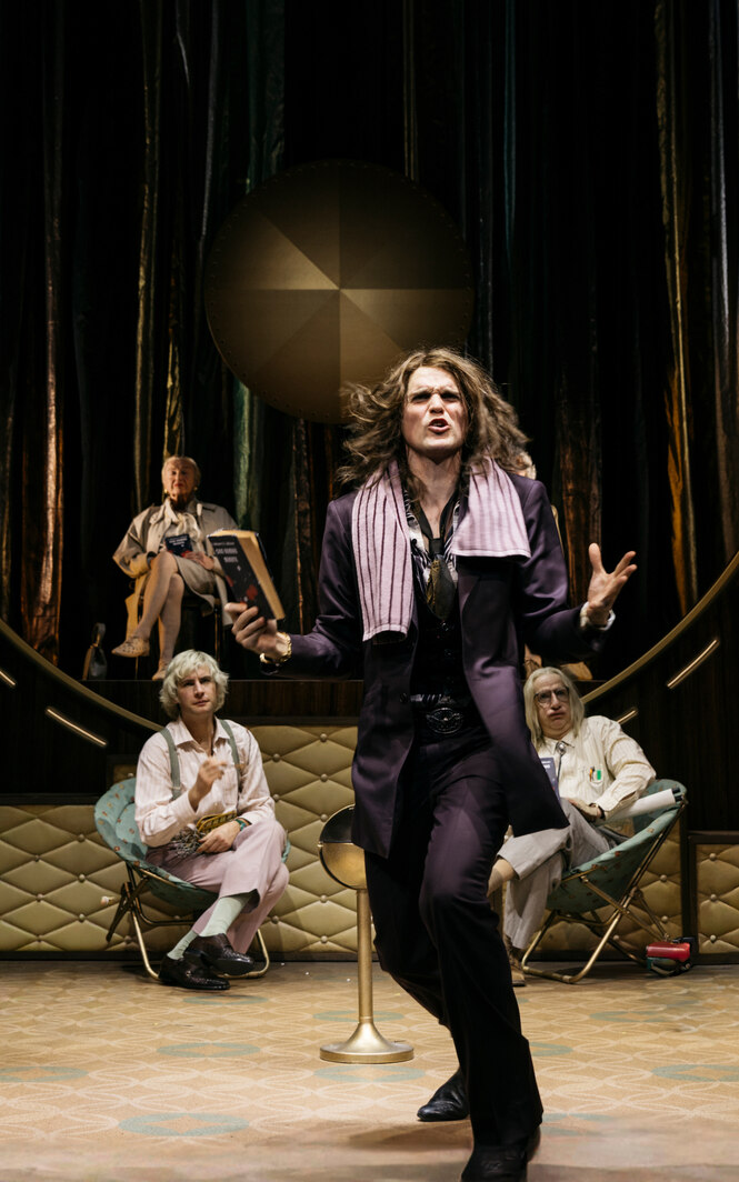 Ein stehender Schauspieler gestikuliert. Hinter ihm sind sitzend drei weitere zu sehen