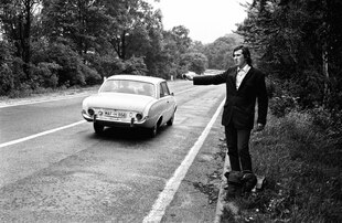 Schwarz-weiß Foto aus den 60er Jahren, ein junger Mann in Anzug und Tasche steht trampend an einer Landstrasse. Ein Auto fährt an ihm vorbei, ohne zu halten