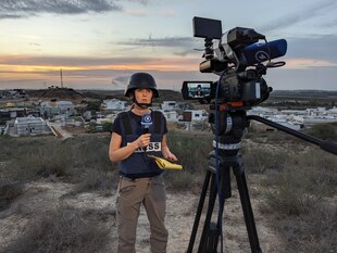 Sophie von der Tann steht mit Schutzweste und -Helm vor einer Siedlung im Abendlicht, im Vordergrund ist eine Fernsehkamera auf sie gerichtet