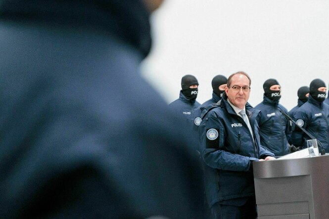 Ein Mann steht an einem Rednerpult, eineige vermummte Personen stehen daneben