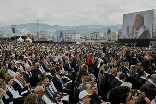Viele Menschen sitzen in Reihen und schauen auf Monitore, die den Papst Leo XIV zeigen