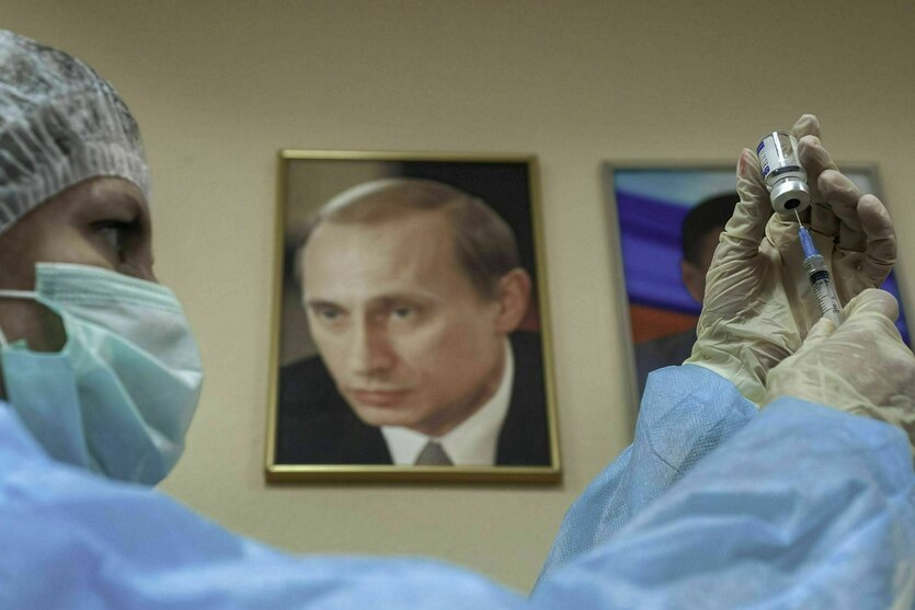 Eine Person zieht eine Spritze auf, an der Wand hängt ein gerahmtes Foto von Vladimir Putin