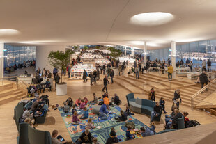 Der Innenraum der öffentlichen Zentralbibliothek „Oodi“ von Helsinki. Zu sehen sind spielende Kinder auf Teppichen, sowie im Hintergrund Bücherregale und große Zimmerpflanzen. Der Raum ist in warmen Licht erleuchtet. Viele Besucher*innen befinden sich in der Bibliothek.