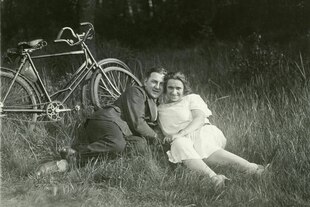 Ein Paar, Mann und Frau liegen im Gras, er trägt Anzug, sie ein helles Kleid und am Baum, der hinter ihnen, steht lehnen zwei Fahrräder