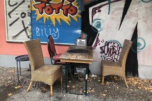 Korbstühle und ein Tisch vor einem Haus, das mit Graffitis bemalt ist, die Rolläden der fenster sind geschlossen