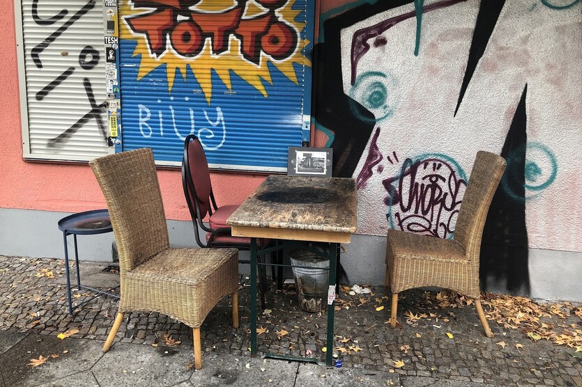 Korbstühle und ein Tisch vor einem Haus, das mit Graffitis bemalt ist, die Rolläden der fenster sind geschlossen