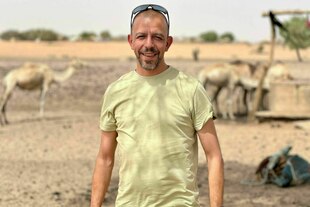 Ein Mann lächelt in die Kamera, hinter ihm stehen Kamele