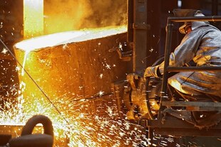 Ein Arbeiter in silbrigem Overall dreht an einem Schwenkrad einen riesigen Bottich gefüllt mit flüssigem Metall, das Funken sprühend in eine stählerne Füllform geschüttet wird.