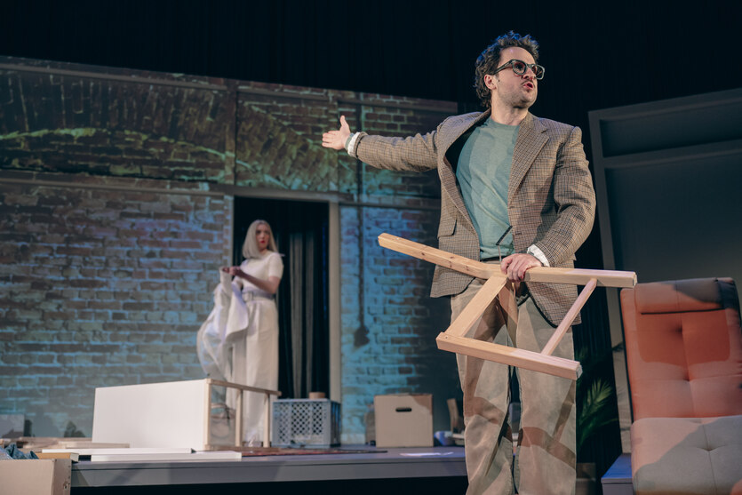 Ein Mann mit einem kaputten Stuhl in der Hand streckt seinen Arm nach hinten, wo eine Frau zwischen zerstörten Möbeln steht