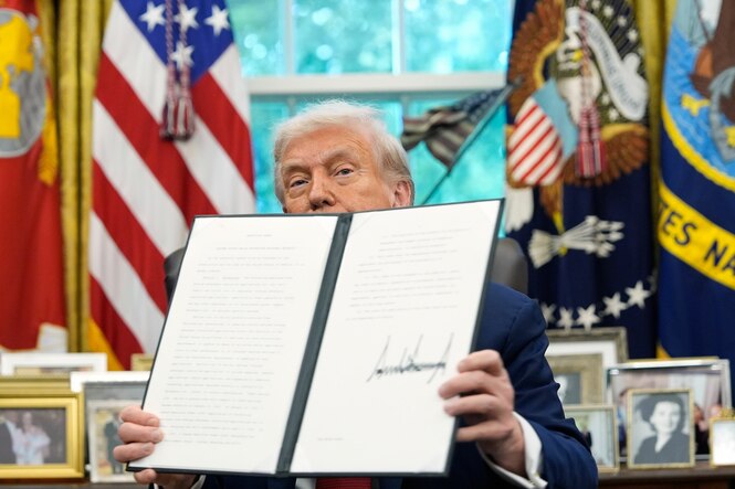 US-Präsident Trump sitzt in seinem Amtszimmer und hält eine große Kladde mit seiner Unterschrift hoch