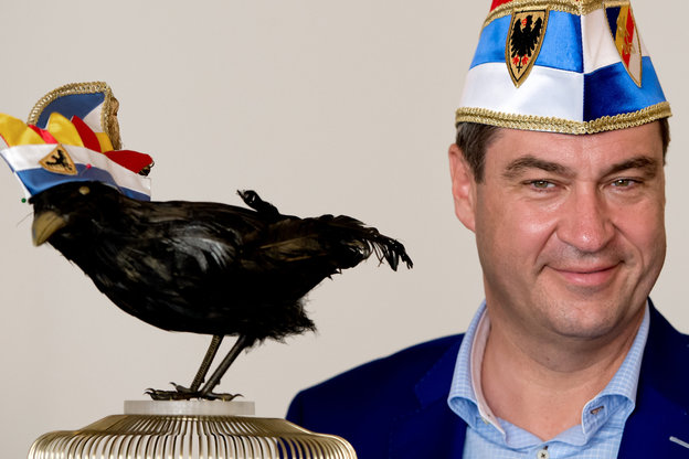 Markus Söder hat eine Narrenkappe auf dem Kopf, vor ihm ein Vogel ebenso mit Narrenkappe