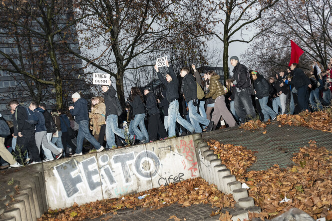 Eine Menge Jugendlicher laufen demonstrierend eine Straße entlang, einige halten Schilder hoch auf denen steht: „Krieg ist dumm“ oder „Nicht Cool“.
