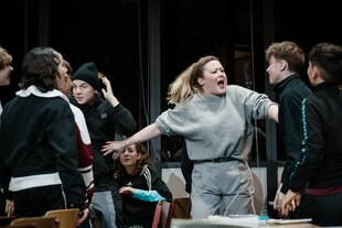Rahel Weiss steht in der Rolle von Carla Nowak gestikulierend inmitten von jungen Menschen, die Schüler darstellen.