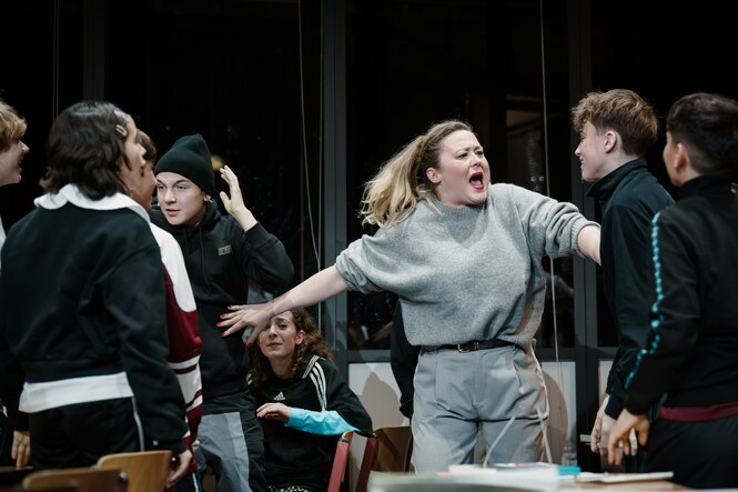 Rahel Weiss steht in der Rolle von Carla Nowak gestikulierend inmitten von jungen Menschen, die Schüler darstellen.