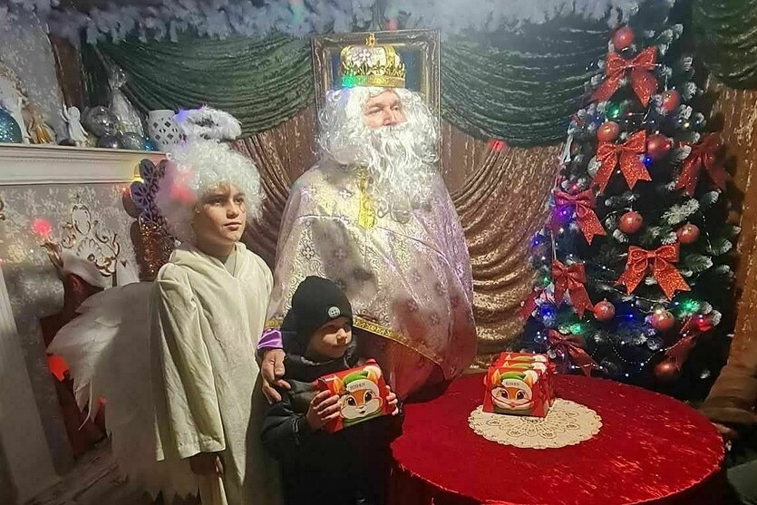 Der Nikolaus in hellem Gewand steht neben zwei Kindern - auf dem Tisch vor ihnen, der Tisch auf dem die Gaben für die Kinder stehen