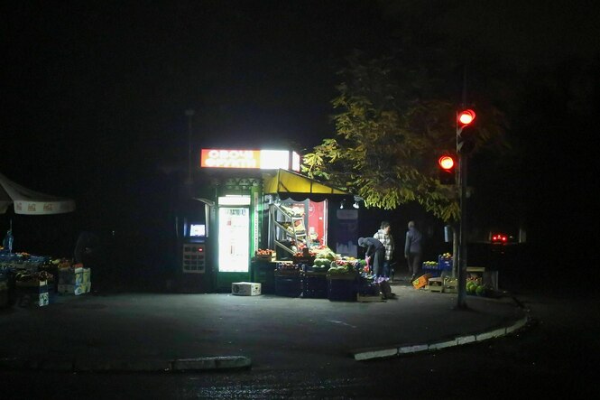Menschen kaufen an einem beleuchteten Obststand ein, der Rest der Straße ist unbeleuchtet