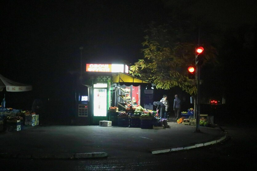 Menschen kaufen an einem beleuchteten Obststand ein, der Rest der Straße ist unbeleuchtet