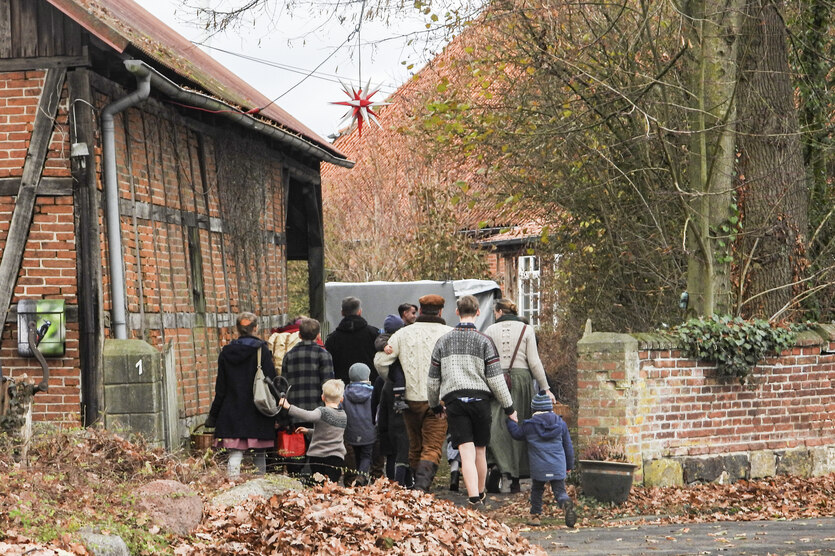 Menschen in Tracht gehen auf einem niedersächsischen Backstein-Fachwerk-Gehöft, sie kehren den Betrachterinnen den Rücken zu