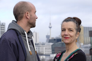 Susanne Memarnia und Erik Peter stehen im Freien mit Blick über Berlin – im Hintergrund ist der Berliner Fernsehturm. Die Person links blickt seitlich weg, die Person rechts schaut schmunzelnd in die Kamera.