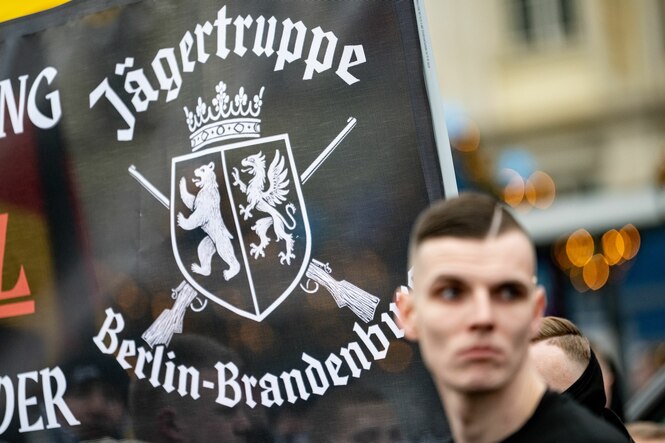Ein Mann mit strammem Seitenscheitel steht vor einem Banner mit der Aufschrift Jägertruppe Berlin-Brandenburg