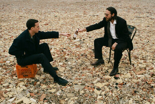 Max Kakacek und Julien Ehrlich der Band Whitney sitzen in Geröll in schwarzer, elegant wirkender Kleidung. Kakacek links auf einem Bierkasten, Ehrlich rechts auf einem Stuhl und reicht Kakacek eine Bierflasche.