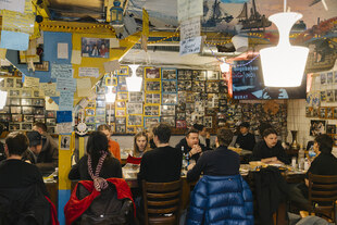 Menschen sitzen um einen großen Tisch herum in einem Gastraum, an den Wänden hängen viele Fotos