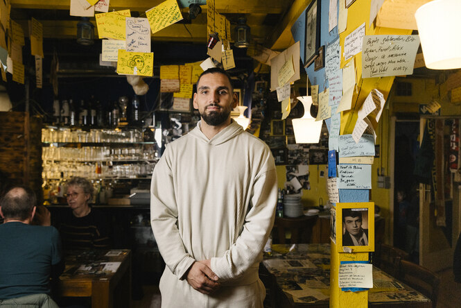 Sezgin Sanar steht in einem Gastraum, erträgt einen cremeweißen Hoodie