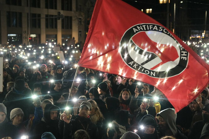 Eine große Menschenmenge bei Dunkelheit, die Demonstrierenden leuchten mit ihren Handys, eine große rote Fahne mit der Aufschrift „Antifaschistische Aktion“ wird geschwungen