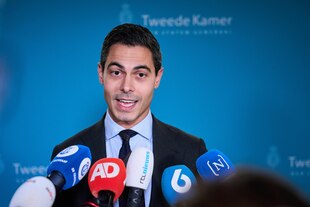 Ein Mann im Anzug vor einer blauen Wand spricht in Mikrofone