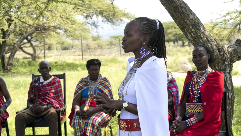 Jacinta spricht zu einer Gruppe, die auf Stühlen unter einem Baum sitzt. Sie trägt ein helles Kleid mit einem Umhang, hochgesteckte Haare, Kette und große Ohrringe