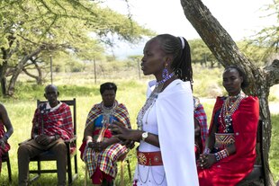Jacinta spricht zu einer Gruppe, die auf Stühlen unter einem Baum sitzt. Sie trägt ein helles Kleid mit einem Umhang, hochgesteckte Haare, Kette und große Ohrringe